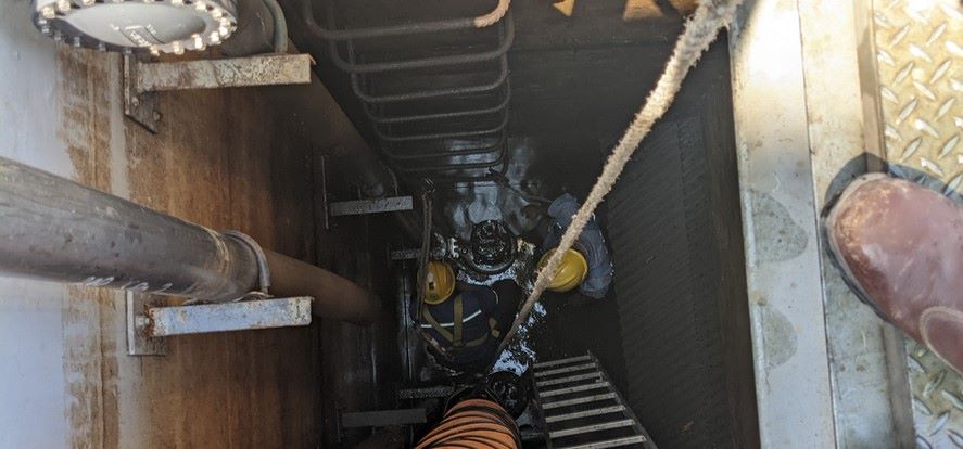 Two workers in safety gear, including helmets, expertly climb down a ladder inside a narrow metal shaft. A rope hangs alongside them amidst various pipes in the confined space, showcasing high-quality civil construction services. A person's boot is partially visible at the edge of the opening.