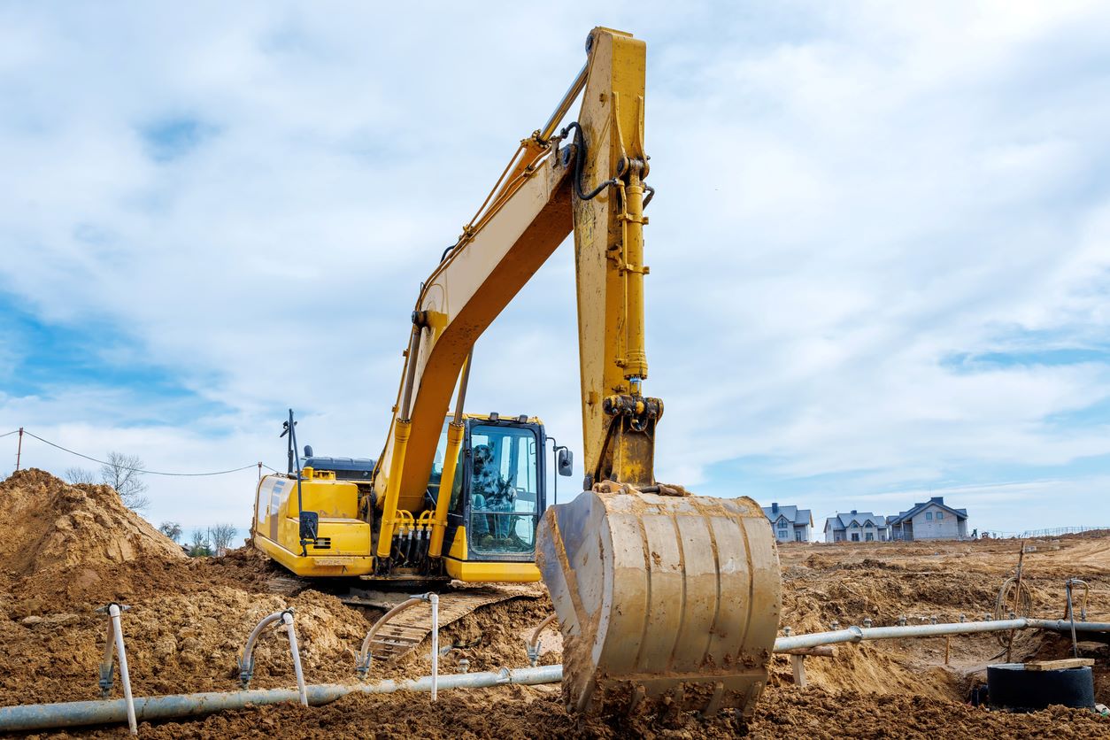 A large yellow excavator is at work on a construction site, showcasing high-quality civil construction services. Its arm is extended, and the bucket is poised over dirt. The scene unfolds under partly cloudy skies with a few buildings visible in the distance.