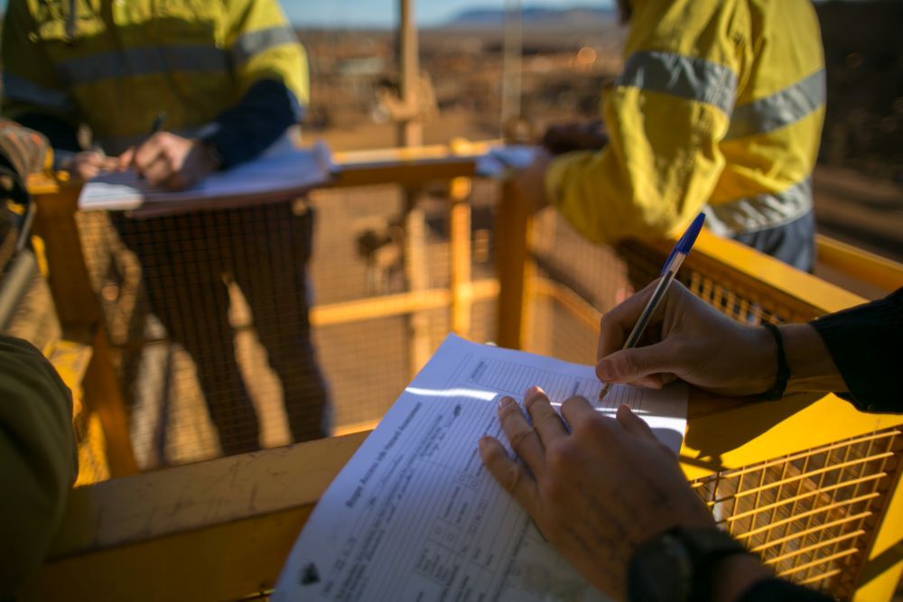 Workers in yellow safety vests, representing high-quality civil construction services, fill out forms on a metal platform with a yellow railing. The scene is outdoors, with blurred industrial equipment in the background and sunlight casting shadows on the papers.