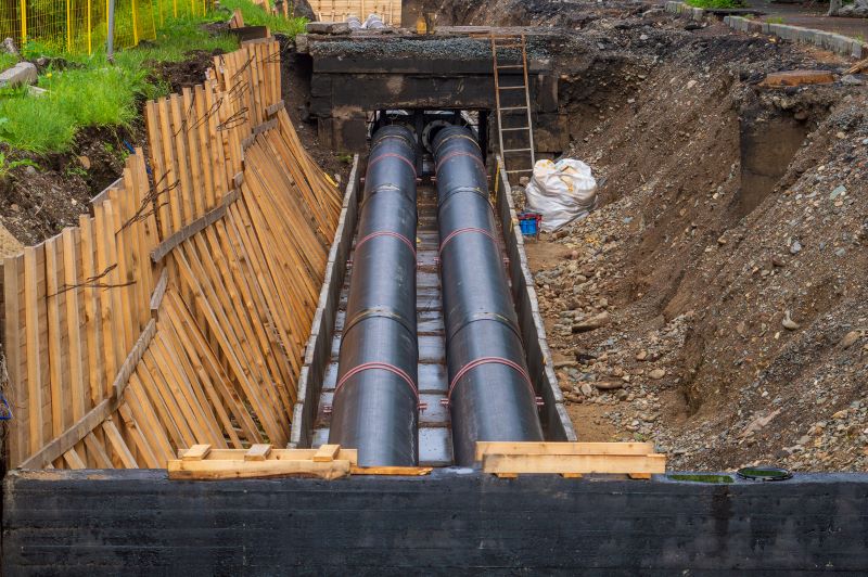 Image of a construction site featuring two large black pipes laid in a trench. Wooden support beams line one side of the trench, which is surrounded by dirt and gravel. A small ladder leans against the trench wall in the background.