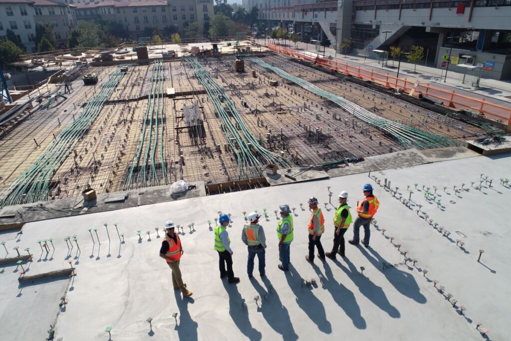 A group of construction workers in safety gear and helmets stand on a concrete surface, reviewing plans. They are on a large construction site with visible wiring and building materials in the background.
