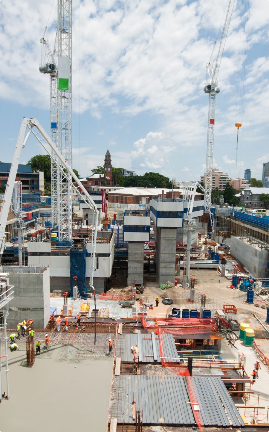 A busy construction site with workers, cranes, and various materials scattered around. Several tall cranes are in operation, and the sky is partly cloudy. In the background, a mix of modern and older buildings is visible.