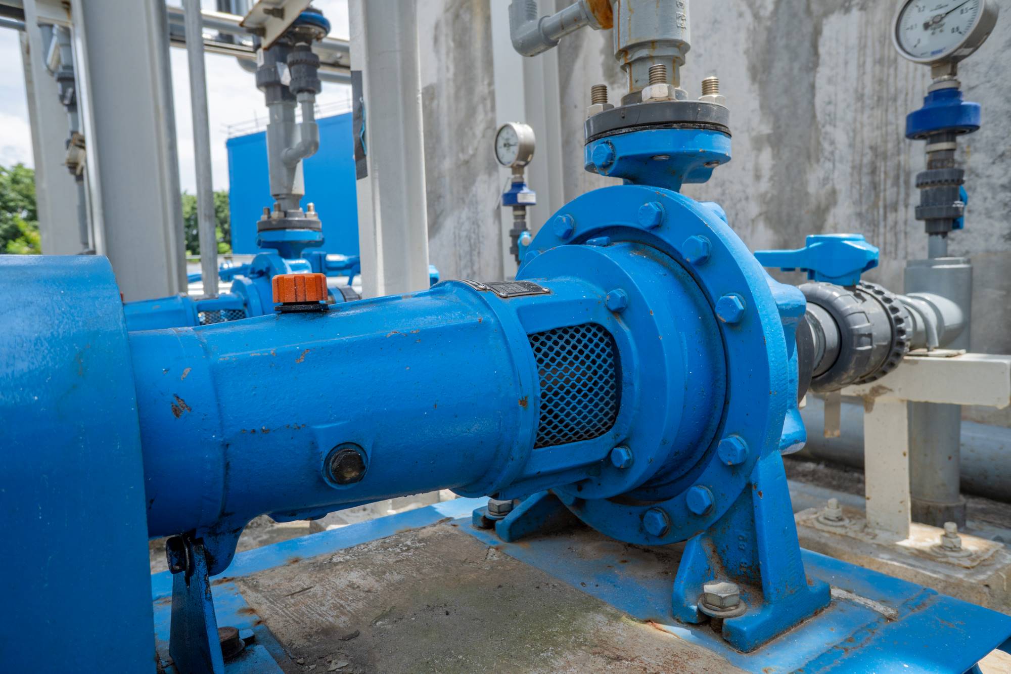 Close-up of a blue industrial water pump with pipes and valves in the background, mounted on a concrete base. Gauges and pipes are visible, indicating a system for controlling fluid flow. The setting is outdoors near a concrete wall.